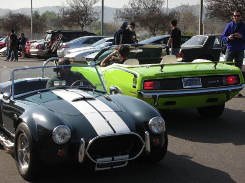 1998 Ford Crown Victoria - dark Shelby Cobra roadster with white racing stripes parks next to a lime-green Plymouth Barracuda at a car meet.