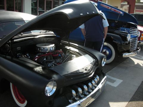 1998 Ford Crown Victoria - matte-black custom hot rod sits with its hood up showing a chromed V8, beside a blue vintage truck at a show.