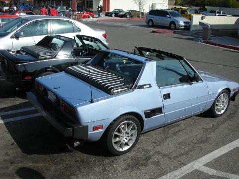 1998 Ford Crown Victoria - light-blue Fiat X1/9 targa with a louvered rear panel parks in a lot with other cars and storefronts behind.