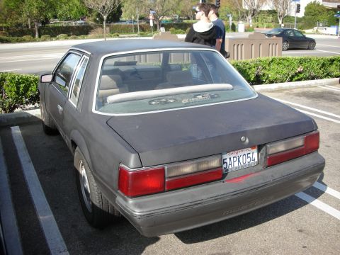 1998 Ford Crown Victoria - Rear three-quarter view of a faded grey 1980s Ford Mustang notchback coupe, plate 5APK054, parked at a meet.