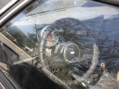 1998 Ford Crown Victoria - Looking through a side window at a worn leather-wrapped steering wheel and dashboard gauges, trees reflected on glass.