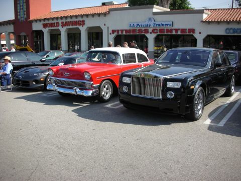 1998 Ford Crown Victoria - black Rolls-Royce Phantom sedan sits beside a red-and-white 1956 Chevrolet and a dark roadster in a shopping-center parking lot.