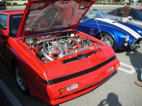 1998 Ford Crown Victoria - red sports car displays its modified twin-turbo engine bay with the hood up, next to a blue racing-striped Cobra-style roadster.