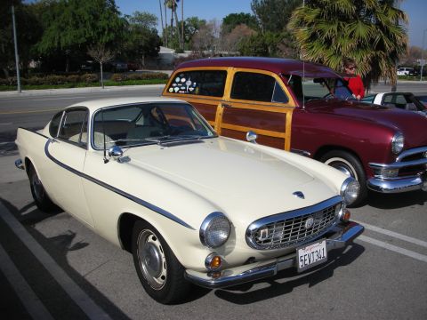 1998 Ford Crown Victoria - cream-colored Volvo P1800 coupe sits in the foreground with a maroon woody-style wagon parked behind it.