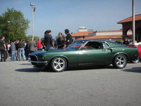 1998 Ford Crown Victoria - dark green late-1960s Ford Mustang fastback sits side-on with chrome bumpers and polished wheels at a busy car show.