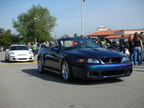 1998 Ford Crown Victoria - blue Ford Mustang SVT Cobra convertible rolls forward past a white Porsche and a crowd at a car gathering.