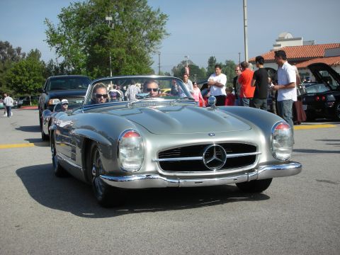 1998 Ford Crown Victoria - silver Mercedes-Benz 300SL roadster with two occupants drives toward the camera through a crowd at a car show.