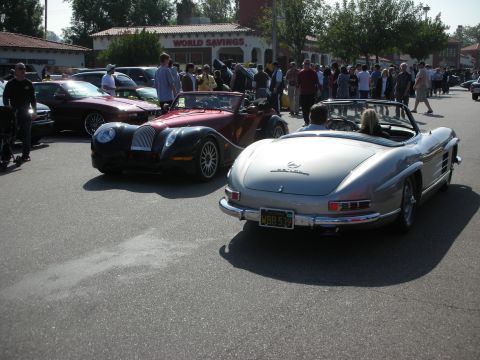 1998 Ford Crown Victoria - silver Mercedes-Benz 300SL roadster is seen from the rear next to a black-and-maroon Morgan-style roadster in a busy lot.