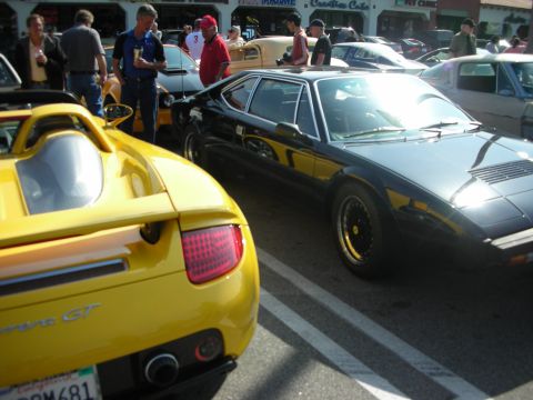 1998 Ford Crown Victoria - yellow Porsche Carrera GT is viewed from the rear next to a black wedge-shaped sports car in a sunlit show lot.