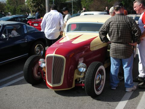 1998 Ford Crown Victoria - red-and-gold vintage hot rod roadster with a chrome grille and whitewall tires draws spectators at a car show.