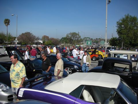 1998 Ford Crown Victoria - Spectators walk among parked sports cars and classics at a sunny outdoor car show, with a purple convertible in the foreground.
