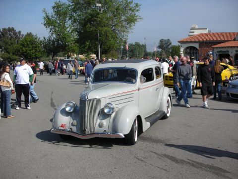 1998 Ford Crown Victoria - white late-1930s Ford street rod two-door sedan rolls slowly through a crowded car show on a sunny day.