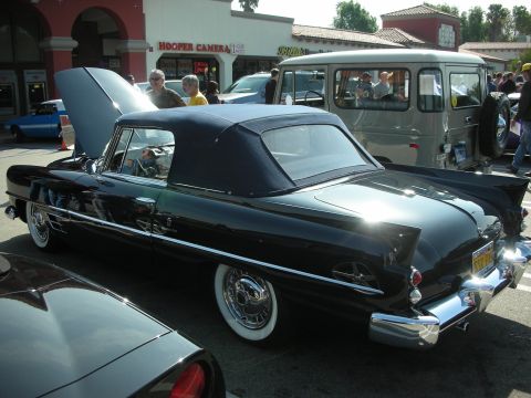1998 Ford Crown Victoria - black late-1940s style convertible with chrome trim and whitewall tires sits parked outside a Hooper Camera storefront.