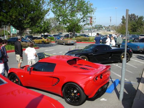 1998 Ford Crown Victoria - red Lotus Elise and a black Lotus park near each other in a lot, with a highway and trees in the background.