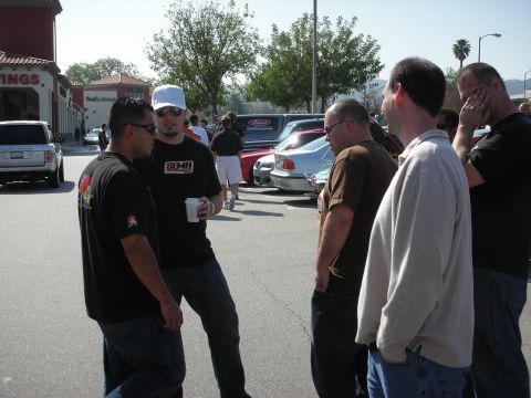 1998 Ford Crown Victoria - group of men in casual clothes stand talking in a parking lot, one wearing a SEMA T-shirt, cars behind them.