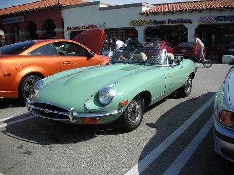 1998 Ford Crown Victoria - mint green Jaguar E-Type roadster with wire wheels parked before a row of shops at a car meet.