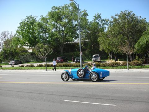 1998 Ford Crown Victoria - blue vintage Bugatti open-wheel race car drives along a tree-lined boulevard with a person walking nearby.