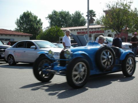 1998 Ford Crown Victoria - blue vintage Bugatti open-wheel race car drives through a lot past a silver sedan and standing spectators.
