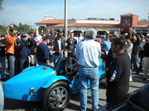 1998 Ford Crown Victoria - dense crowd mingles at a car gathering, including a woman wearing a Ferrari leather jacket near a blue car.