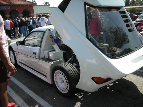 1998 Ford Crown Victoria - white wedge-shaped supercar shows off its turbocharged engine with the rear clamshell bodywork raised at a crowded outdoor car meet.