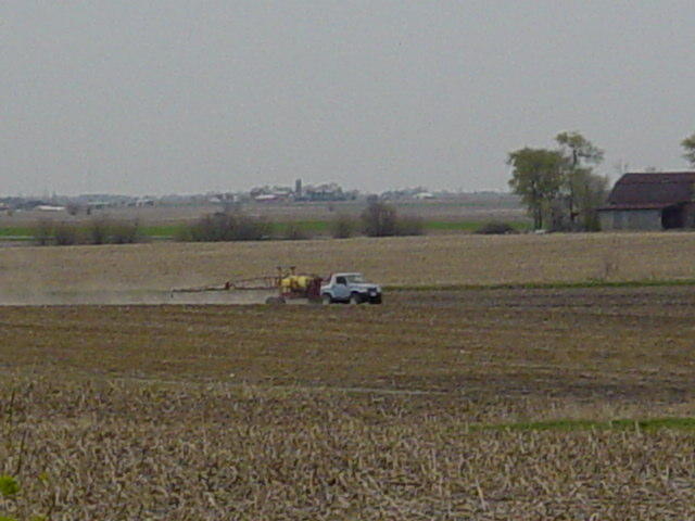 Pickup truck pulling a yellow sprayer across a tilled farm field, town and barn on the horizon