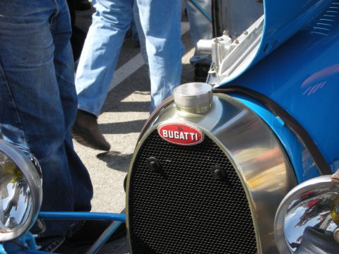 1998 Ford Crown Victoria - Close-up of the polished horseshoe radiator shell and red oval Bugatti badge on a blue vintage racer, with onlookers behind.