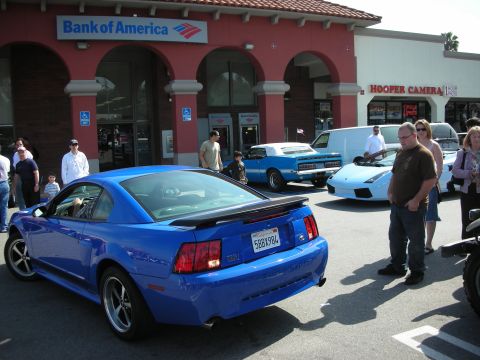 1998 Ford Crown Victoria - blue Ford Mustang coupe parked at an outdoor car meet in front of a Bank of America and Hooper Camera storefront.