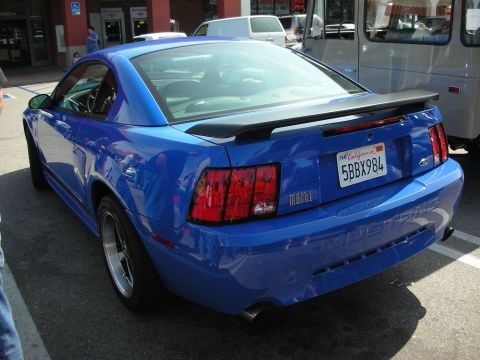 1998 Ford Crown Victoria - blue Ford Mustang Mach 1 coupe with rear spoiler and California plate seen from the rear three-quarter in a lot.