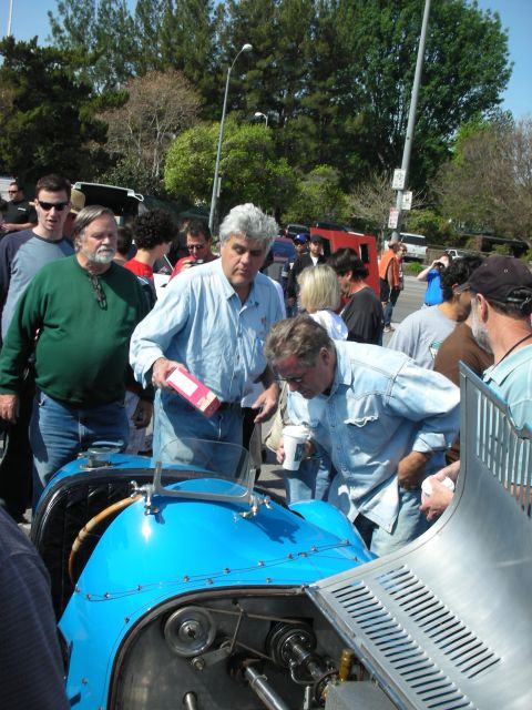 1998 Ford Crown Victoria - gray-haired man holding a pink box leans over a blue vintage race car as onlookers gather at an outdoor event.
