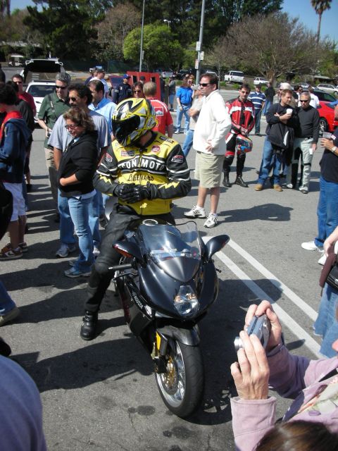 1998 Ford Crown Victoria - rider in yellow leathers and a helmet sits on a black sportbike amid a crowd of spectators in a parking lot.