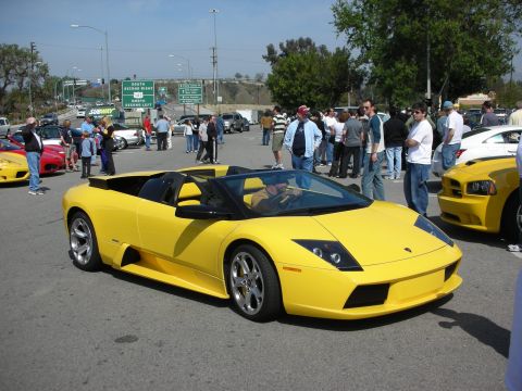 1998 Ford Crown Victoria - yellow Lamborghini Murcielago roadster rolls through a crowded outdoor car show with spectators and freeway signs behind.