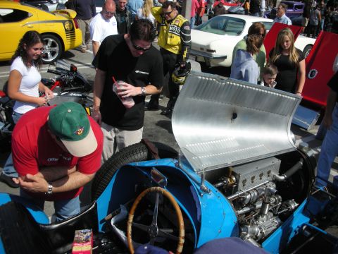 1998 Ford Crown Victoria - man in a red shirt and others crowd around a blue vintage race car with its aluminum hood raised at a street show.