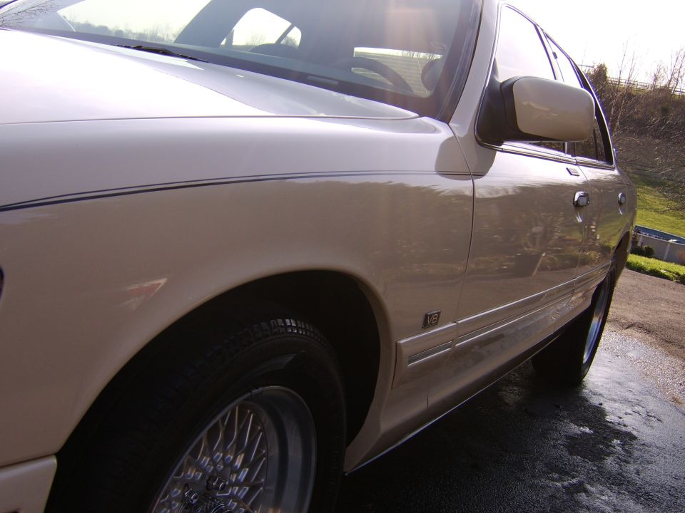 Low side view of tan sedan showing V8 badge, chrome wheel and rear door on wet pavement