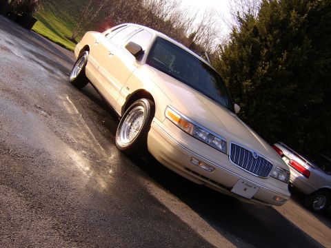 1996 Mercury Grand Marquis - A tan sedan with wire wheels is parked on a wet asphalt driveway with trees in the background.
