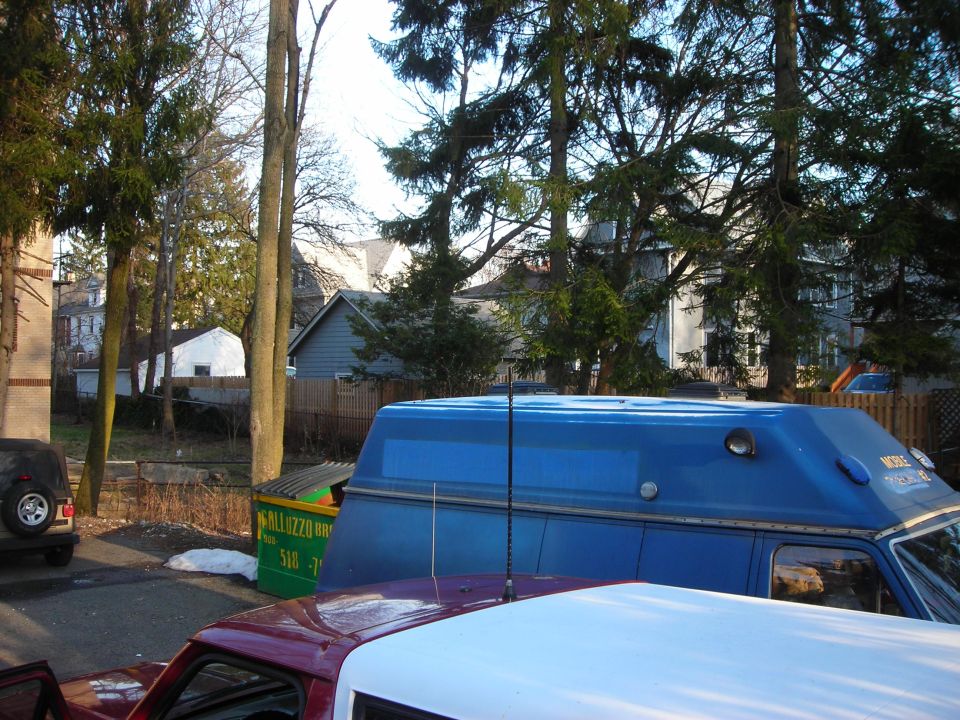 Blue van with a raised roof and antenna, parked next to a dumpster and a red truck