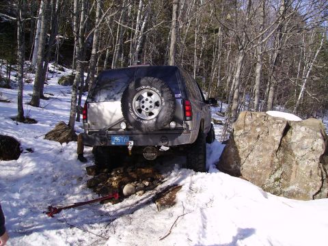 2000 Chevrolet Tahoe - A lifted SUV with a spare tire on the back navigates snowy, rocky terrain, with its license plate visible.