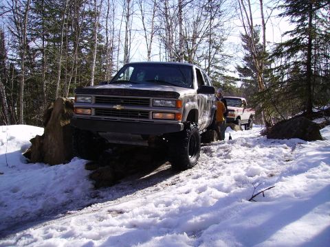 2000 Chevrolet Tahoe - Two SUVs tackle a snow-covered trail in a wooded area.