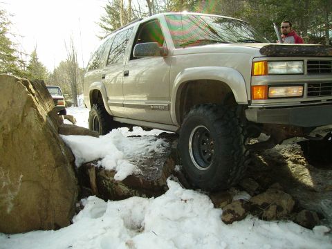 2000 Chevrolet Tahoe - A tan SUV with aggressive tires is shown off-roading over rocks and snow, with another vehicle in the distance.