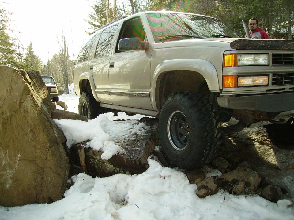 Tan SUV with lifted suspension navigating rocky, snowy terrain. Another vehicle is visible in the background
