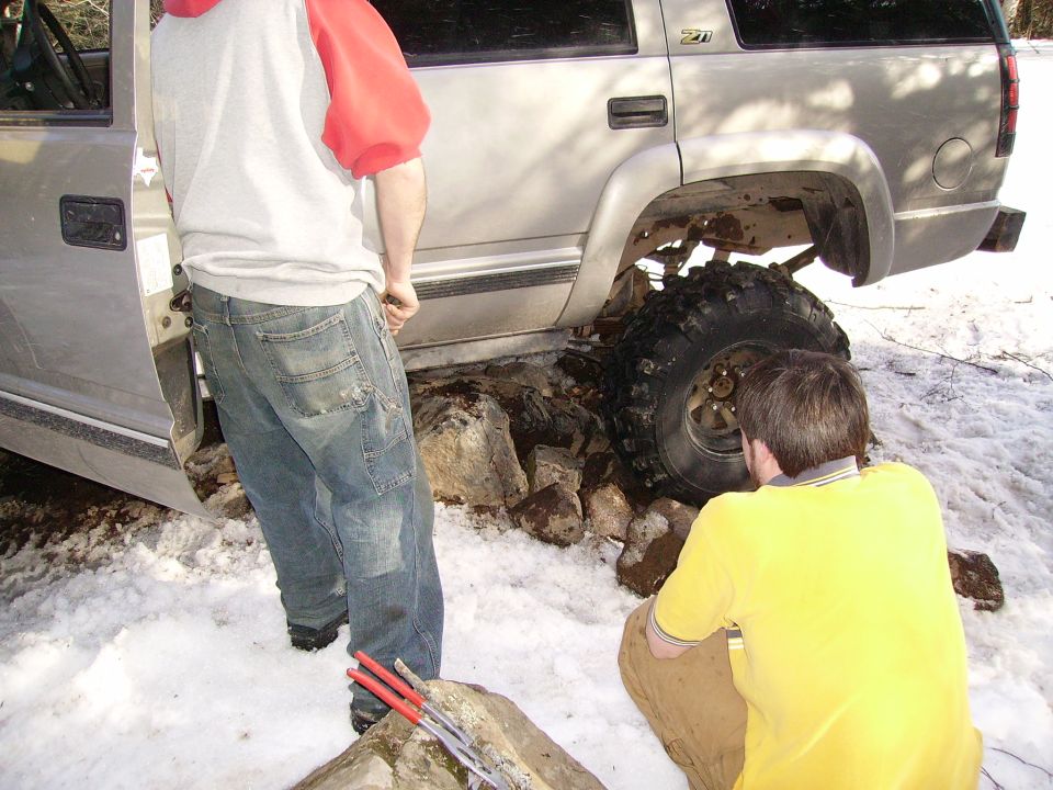 Silver SUV with one wheel stuck on rocks in snow. Two people are looking at it