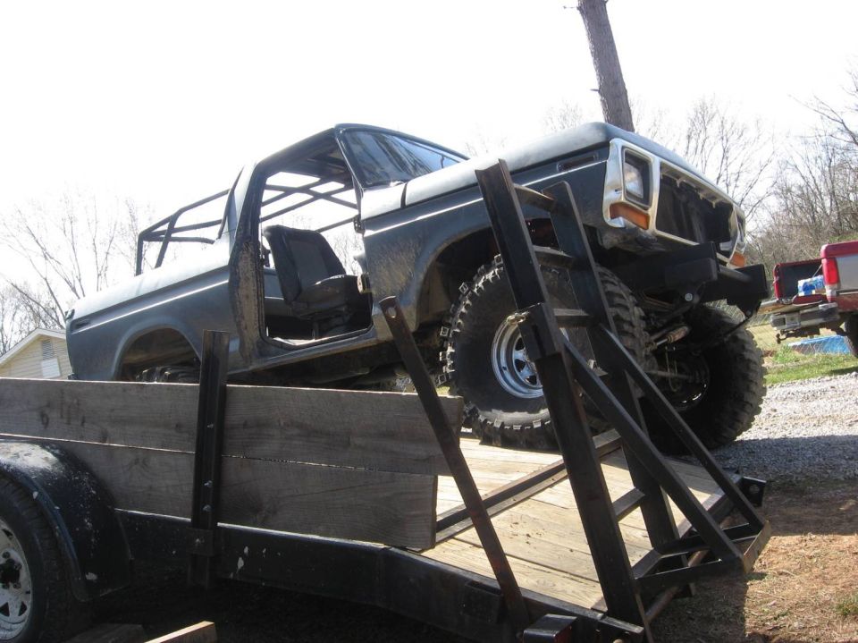 Old Ford Bronco on a trailer, looks like it's being hauled somewhere. It has a roll cage inside and big off-road tires