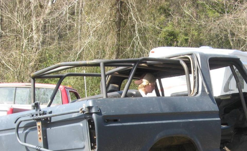 Side view of a dark-colored SUV with a roll cage, a person wearing a hat inside. Another red truck is visible behind it