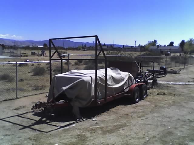 Red tandem-axle utility trailer with tarp-covered load and metal frame, parked on dirt lot