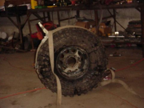 1992 Ford Bronco - A dirty spare tire and wheel is secured with straps in a workshop environment.