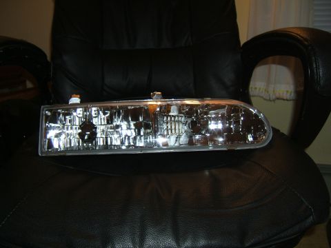 1998 Ford Taurus - A single headlight assembly is shown resting on a black leather chair, with a watermark for Supermotors.net at the bottom right.