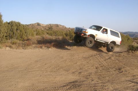 1996 Ford Bronco - A white SUV navigates a dusty off-road trail, its tires kicking up dirt as it climbs a hill.