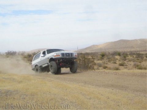 1996 Ford Bronco - A modified Ford Bronco drives off-road, its tires kicking up dust in a desert environment.