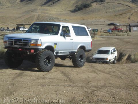 1996 Ford Bronco - Two white SUVs, one heavily modified, drive on a dirt track in a dry, hilly area.