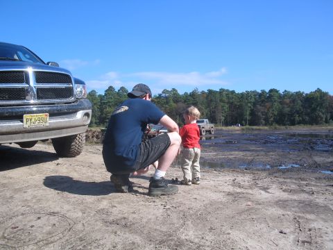 1990 Ford Bronco - Man and child observe trucks in a muddy outdoor area under a clear blue sky.