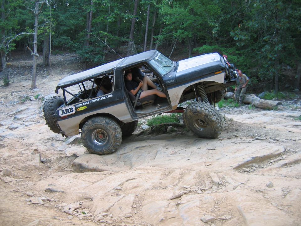 Modified Ford Bronco with large tires and roll cage on rocky terrain. ARB and HIGHANGLEDRIVELINE.COM decals visible